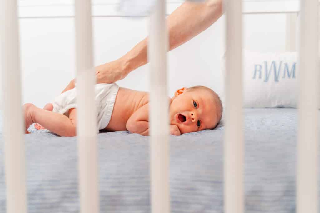 Baby boy yawning, laying crib during lifestyle newborn photography session.