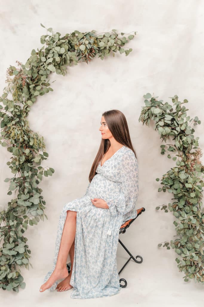 Pregnant woman sitting on stool in front of hand painted backdrop and fresh eucalyptus garland during Chattanooga TN maternity style session