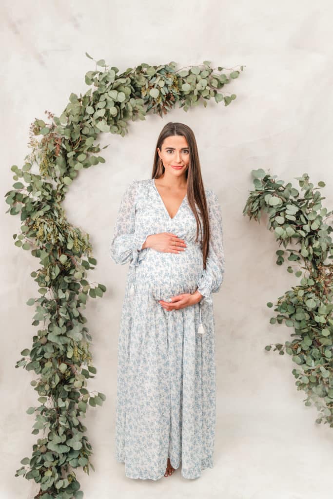 Mother poses under eucalyptus garland during styled maternity photography session with Chattanooga photographer who offers maternity and newborn photo packages. Blue Floral Maternity Gown.