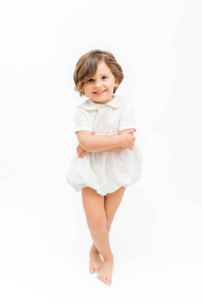 Preschool boy posing with arms and legs crossed during heirloom portrait session in front of white backdrop