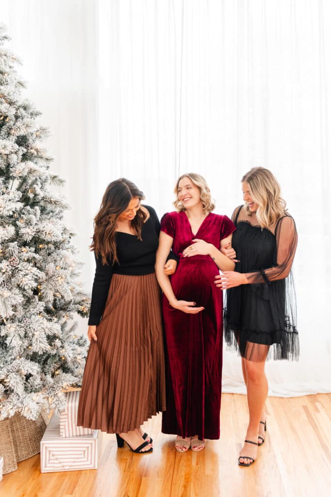Models laugh in front of Christmas tree wearing feminine and bump friendly dresses