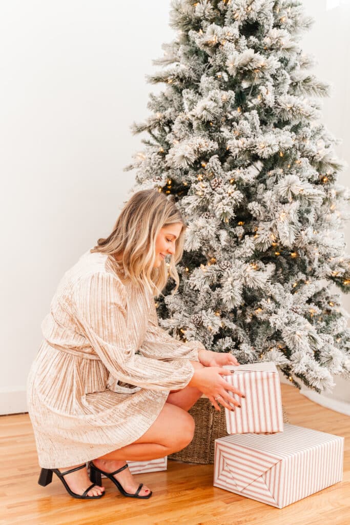 Model holds present in front of Christmas tree wearing shimmery cream dress.