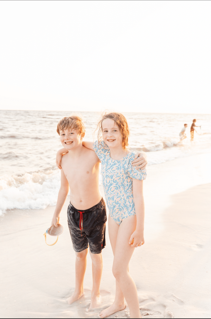 Two kids smile on the beach for photo, but there are people in the background. Post about how to edit out someone in a photo using photoshop.