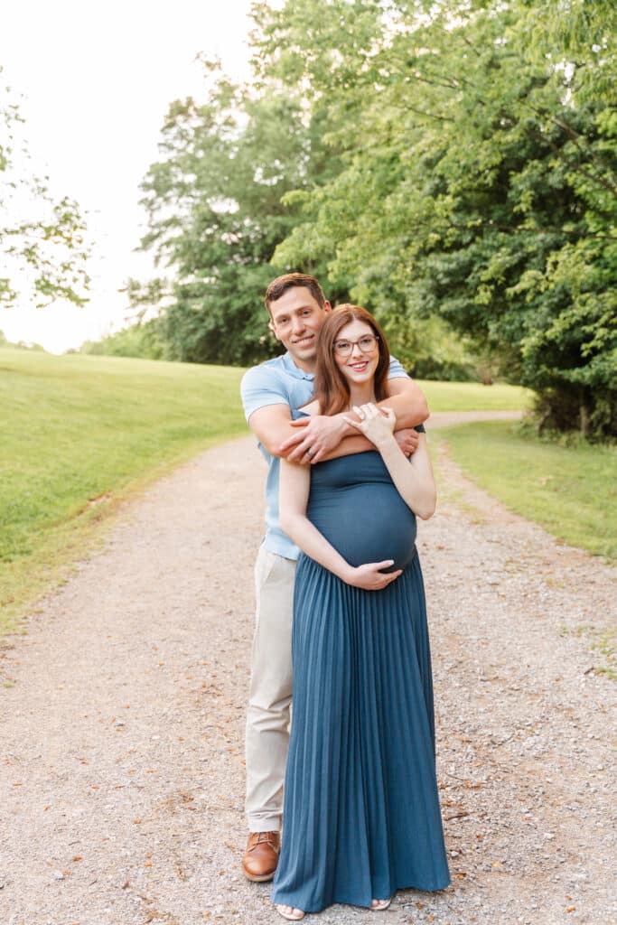 Wondering when to take maternity photos? Chattanooga maternity photographer recommends between 28-34 weeks.  Image of expecting mother and her husband posing together on gravel path in greenway area.  Expecting mother is wearing long blue dress. 