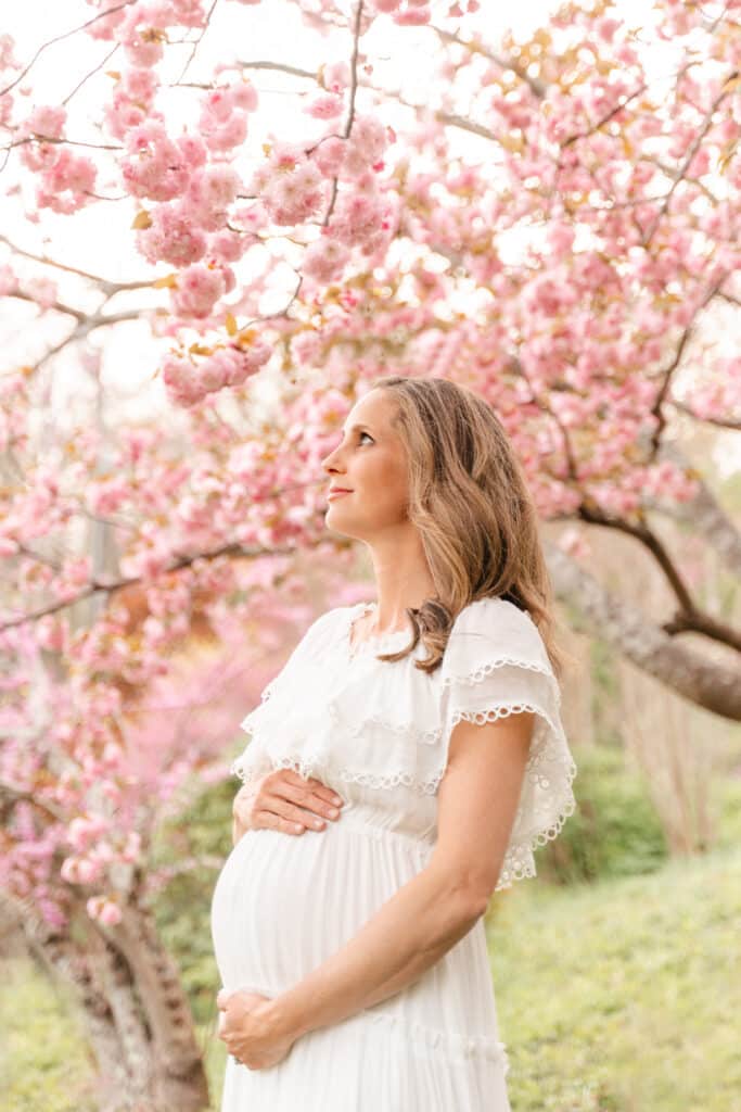 Maternity portrait of expecting mother standing in front of pink cherry blossoms. This expecting mother was photographed at 26 weeks with her 4th child.